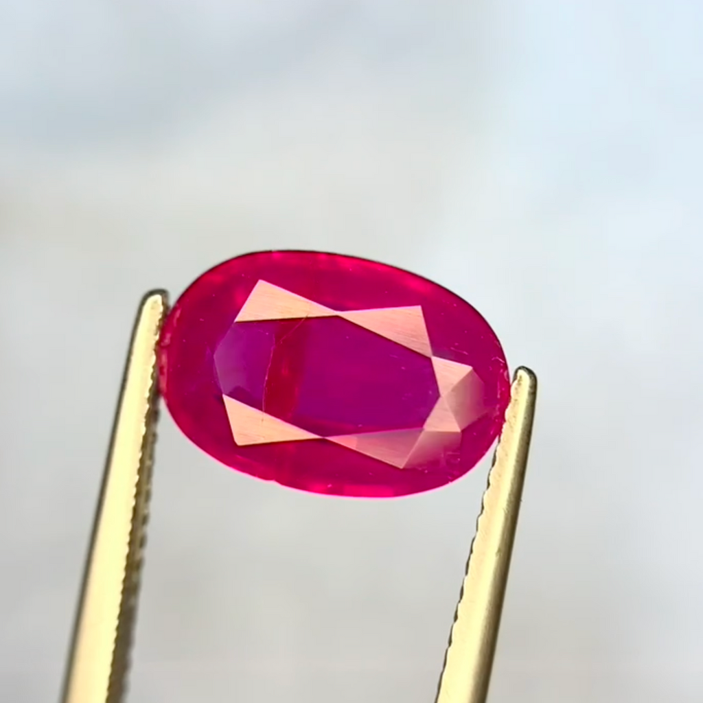Oval-shaped pink gemstone held between gold tweezers on a light gray background